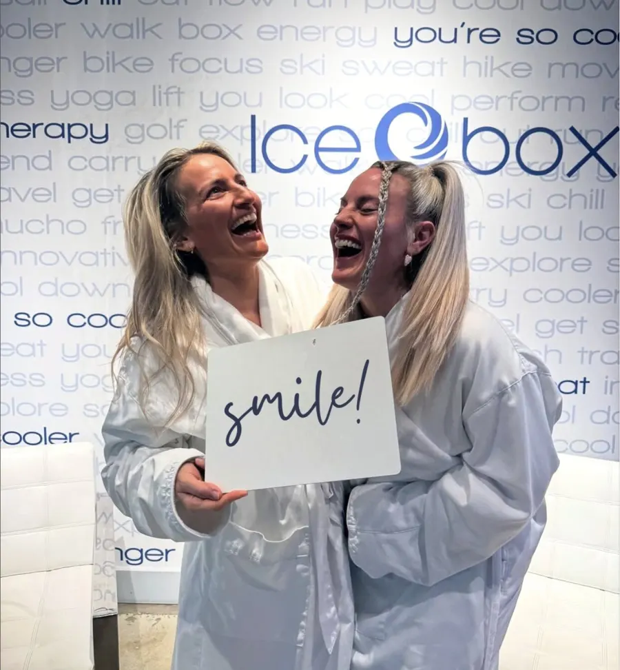 Two friends in white robes laughing together and holding a smile! sign in front of the Icebox wall